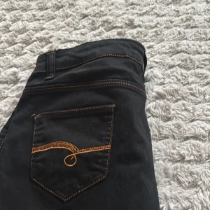 Svarta jeansshorts med kontrastsömmar - Säljer ett par svarta jeansshorts med rå kant och snygga kontrastsömmar i orange. Shortsen har två bakfickor med unik broderad detalj och bälteshällor. Perfekta för dig som vill ha en cool och avslappnad stil.