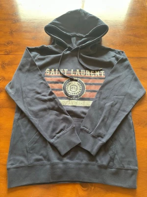 Mörkblå hoodie från Saint Laurent - Mörkblå hoodie från Saint Laurent med tryck i vitt och rött framtill. Klassisk huva med dragsko och känguruficka. Trycket har texten 'SAINT LAURENT' och ett emblem med SL och stjärnor. Tillverkad i mjukt bomullsmaterial, perfekt för en avslappnad look.