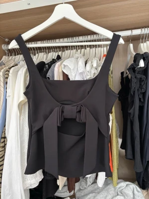 Svart topp med rosettdetalj från Zara - Trendig svart topp med breda axelband och en stor rosett framtill. Toppen har en snygg cut-out under rosetten och en rak, något figurnära passform. Perfekt att styla med jeans eller kjol för en cool look🖤säljer pågrund av att den inte kommit till användning!