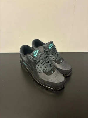 Nike Air Max 90 svart/turkos sneakers - Nike Air Max 90 sneakers i svart mesh och syntet med turkosa detaljer på plös, häl och sidor. Klassisk låg siluett med synlig Air-enhet i sulan och ikonisk Swoosh på sidan. Sköna och stilrena skor som passar perfekt till streetwear.