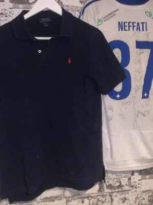 Mörkblå piké från Ralph Lauren - Klassisk mörkblå pikétröja från Polo Ralph Lauren med röd broderad logga på bröstet. Tröjan har korta ärmar, krage och två knappar vid halsen. Tillverkad i bomull för en bekväm och stilren look.