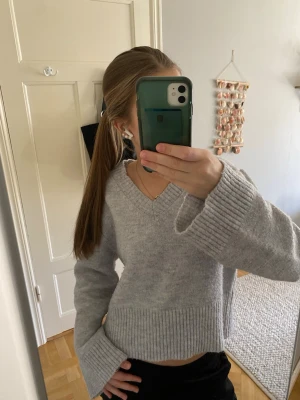 Grå stickad tröja V-ringad - Fin ljusgrå stickad tröja med V-ringning och breda ribbade muddar vid ärmslut och nederkant. Modellen har en avslappnad passform med vida ärmar.