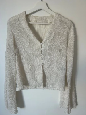 Vit plisserad blus med knappar - Trendig vit blus med plisserat tyg och dekorativa knappar framtill. Blusen har v-ringning och långa ärmar med lätt utsvängd form. Perfekt för dig som gillar en stilren och elegant look.