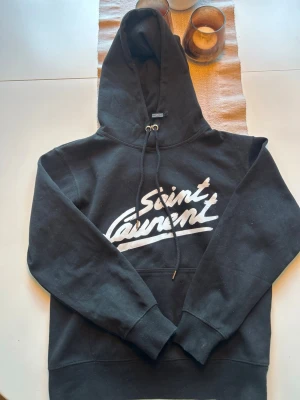 Svart Saint Laurent hoodie med tryck - Snygg svart hoodie från Saint Laurent med stort vitt logotyp-tryck på bröstet. Klassisk modell med huva och dragsko samt ribbade muddar vid ärmar och nederkant. Tillverkad i Italien, mjukt och bekvämt material som passar perfekt till streetwear-stilen.