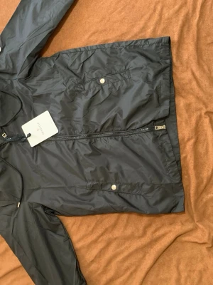 Svart vindjacka från Moncler M - Snygg svart vindjacka från Moncler med dragkedja framtill och två fickor med tryckknappar. Jackan har en minimalistisk design och är tillverkad i ett lätt, glansigt material som står emot vind. Perfekt för dig som vill ha en stilren och funktionell jacka.