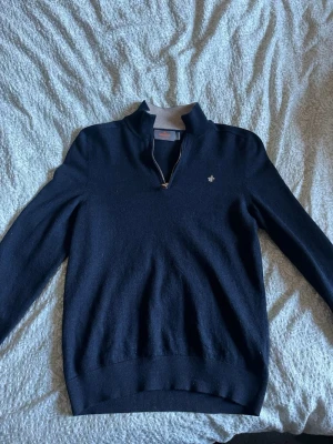 Mörkblå stickad tröja med halvzip - Snygg mörkblå stickad tröja med halvzip och liten broderad logga på bröstet. Tröjan har hög krage och ribbade muddar vid ärmslut och nederkant. Perfekt för lager på lager och enkel att matcha med jeans eller chinos.