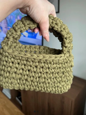 Olivgrön virkad handväska - Unik handväska i olivgrönt, grovt virkat garn. Väskan har en rektangulär form med rundade kanter och ett matchande handtag. Perfekt accessoar för dig som gillar handgjorda detaljer och trendig design.