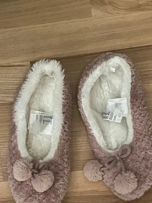 Rosa fluffiga tofflor med bollar H&M - Mysiga rosa tofflor från H&M med fluffig utsida och vit mjuk insida. Dekorerade med två stora bollar framtill för en gullig look. Perfekta att glida runt i hemma när du vill ha det extra bekvämt och varmt.