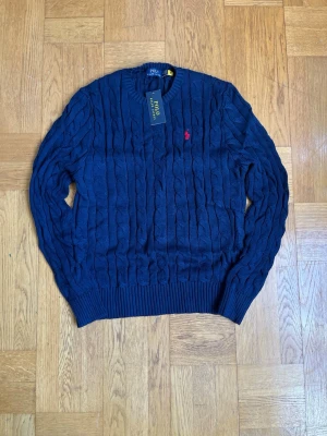 Mörkblå kabelstickad tröja Polo Ralph Lauren - Kabelstickad mörkblå tröja från Polo Ralph Lauren med rund halsringning och klassisk röd logga broderad på bröstet. Tröjan har ribbade muddar vid ärmslut och nederkant. Perfekt för dig som gillar tidlös och stilren design.