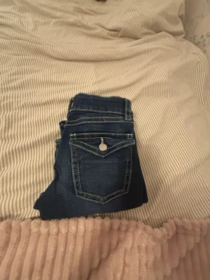 Mörkblå jeans med kontrastsömmar - Säljer ett par mörkblå jeans med vita kontrastsömmar och snygga detaljer på bakfickan. Jeansen har klassisk femficksdesign och silverfärgad knapp. Perfekta till en avslappnad stil och passar till det mesta i garderoben. Finns lite lösa trådar som vissas på bilden och pris kan diskuteras och storlek 152