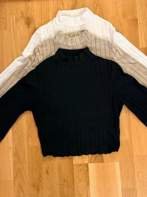 3 stickade polotröjor i olika färger - Säljer tre ribbstickade polotröjor med lång ärm i svart, beige och vitt. Alla har en mellanhög krage och är tillverkade i mjukt, stretchigt material. Perfekta att matcha med jeans eller kjol för en clean look.