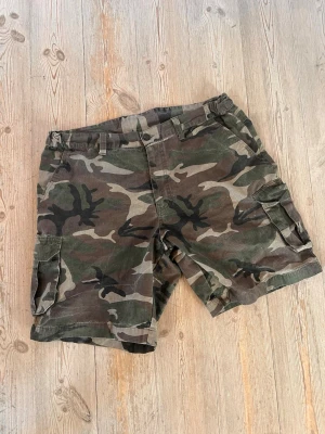 Camo cargoshorts  - Säljer ett par klassiska cargoshorts i camouflage med flera praktiska fickor på sidorna. Shortsen har bälteshällor och stängs med knapp och dragkedja. Swag