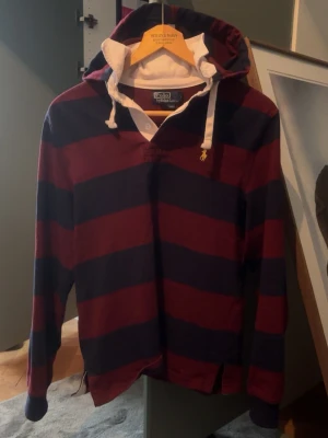 Rugby hoodie vintage Polo Ralph Lauren - Snygg hoodie från Polo Ralph Lauren med breda marinblå och vinröda ränder. Klassisk huva med vita snören och vit insida, samt gul broderad logga på bröstet. Tillverkad i mjuk bomull och har långa ärmar med muddar. Bröstmått tvärs över: 46 cm, längd: 63 cm 