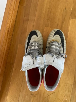 Nike Mercurial vita fotbollsskor - Säljer ett par Nike Mercurial fotbollsskor i vitt med svarta och röda detaljer. Skorna har klassisk snörning, låg profil och dobbar för spel på gräs. Swoosh-loggan syns tydligt på ovansidan och sidan. Perfekta för dig som vill ha lätta och snabba skor på planen.
