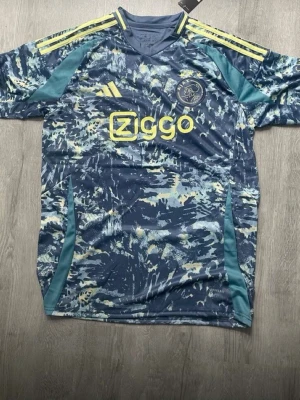 Ajax Jersey  - Snygg blå Ajax matchtröja från Adidas med gult Ziggo-tryck och klubbmärke på bröstet. Tröjan har ett unikt abstrakt mönster i blått, vitt och gult samt gula ränder på axlarna. Tillverkad i lätt och ventilerande funktionsmaterial, perfekt för fotbollsträning.