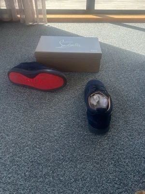 Mörkblå sneakers från Christian Louboutin - Snygga mörkblå sneakers från Christian Louboutin med klassisk röd sula och snörning. Skorna är tillverkade i mocka och har en stilren design med diskreta sömmar och beige insida. Perfekta för dig som vill ha något exklusivt och trendigt.