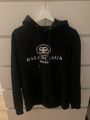 Svart hoodie med tryck - Snygg svart hoodie med vit logga och text på bröstet. Klassisk huva med dragsnören och stor magficka framtill. Perfekt för dig som gillar streetwear och vill ha en exklusiv vibe. Mjuk och skön bomullskänsla.