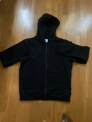 Svart zip hoodie med korta ärmar - Säljer en svart hoodie med dragkedja framtill och korta ärmar. Hoodien har en stor huva med dragsko och en klassisk magficka. Perfekt för en avslappnad streetwear-look. Materialet känns mjukt och bekvämt, troligen bomull eller bomullsblandning.