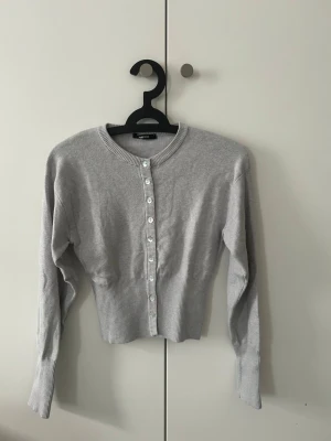 Ljusgrå kofta från Gina Tricot - Ljusgrå ribbstickad kofta från Gina Tricot med rund halsringning och pärlemorknappar framtill. Kortare modell med långa ärmar och bred ribbad kant nedtill och vid ärmsluten. Perfekt att styla över ett linne eller t-shirt. Ny utan prislapp