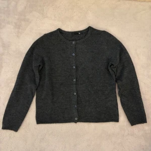 Mörkgrå basic kofta med knappar - En enkel och stilren mörkgrå kofta med rund halsringning och knäppning framtill. Koftan har långa ärmar och är tillverkad i ett mjukt stickat material. Perfekt att slänga över en t-shirt eller linne för en avslappnad look.