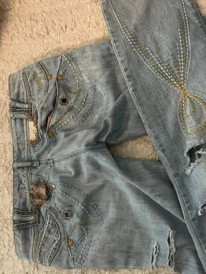 Snygga jeans med detaljer - Unika ljusblå jeans med snygga slitningar, dekorativa broderier i gult och vitt samt detaljerade fickor. Jeansen har trasiga partier och coola knappar i olika färger. Perfekta för dig som vill sticka ut med din stil.