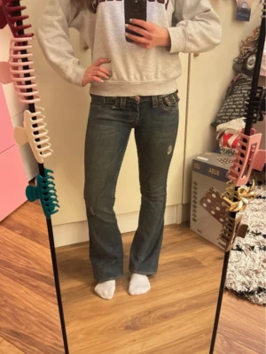 True Religion bootcut jeans  - Säljer ett assnygga par blå bootcut jeans från True Religion med snygga slitningar och detaljerade bakfickor med lock och knapp. Klassisk femficksmodell i denim med låg midja och kontrastsömmar. Perfekta för en avslappnad och trendig look.