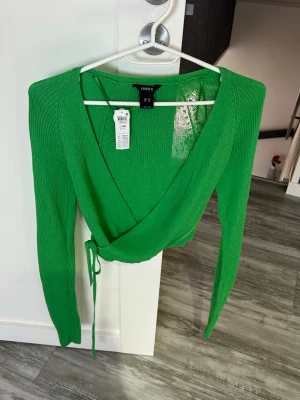 Grön omlottopp från Lindex - Snygg grön omlottopp från Lindex med långa ärmar och ribbad struktur. Tröjan har knytband i midjan och djup v-ringning, perfekt för att skapa en trendig lager-på-lager-look. Materialet är mjukt och stretchigt för skön passform.