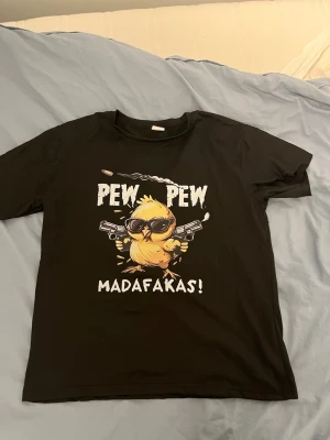 Svart t-shirt med kycklingtryck - Svart t-shirt med ett stort tryck av en gul kyckling med solglasögon och två pistoler samt texten 'PEW PEW MADAFKAS!' på framsidan. T-shirten har rund hals och korta ärmar. Perfekt för dig som gillar humoristiska motiv.