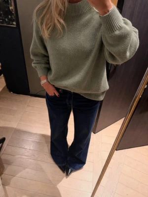 Grön stickad tröja  - Mysig stickad tröja i dämpad grön färg med bred ribbad mudd vid hals, ärmslut och nederkant. Tröjan har ballongärmar och en avslappnad passform. Perfekt att styla med jeans eller kjol för en trendig look.