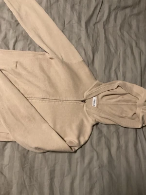 Beige hoodie med dragkedja från RYVLS - Snygg beige hoodie från RYVLS Stockholm med dragkedja framtill och huva. Tillverkad i mjukt material som känns skönt mot huden. Perfekt för en avslappnad och stilren look. Passar dig som gillar enkel och clean stil.