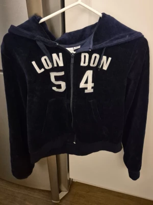 Mörkblå hoodie med dragkedja - Mysig mörkblå hoodie i mjuk velour material med huva och dragkedja framtill och fickor. På bröstet står det 'LONDON 54' i vita bokstäver och siffror. Hoodien har långa ärmar och ribbade muddar vid ärmslut och nederkant. Perfekt för en avslappnad stil.