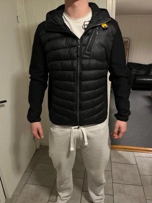 Svart cardigan från Parajumpers - Snygg svart cardigan från Parajumpers med huva och dragkedja framtill. Jackan har quiltad front, släta ärmar och en gul detalj vid halsen. Märkets patch sitter på vänster ärm. Perfekt för kalla dagar.