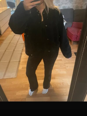 Svart teddyjacka med tryckknappar - Mysig svart teddyjacka med oversized passform och rund hals. Jackan har tryckknappar framtill och är gjord i fluffigt teddy-material som ger en trendig look. Perfekt för dig som vill ha en stilren och enkel jacka till hösten eller vintern. En knapp på fickan har lossnat. 