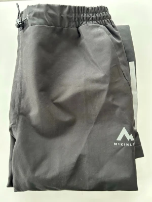 Svarta täckbyxor från McKinley - Säljer ett par svarta täckbyxor från McKinley med elastisk midja och dragsko. Byxorna har en enkel, stilren design och är perfekta för kalla dagar. Materialet är slitstarkt och passar bra för utomhusaktiviteter.