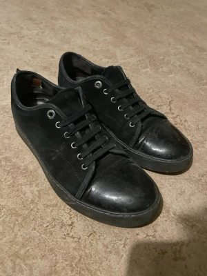 Lanvins helsvarta  - Säljer ett par helsvarta sneakers med lågt skaft och snygga mockadetaljer på sidorna. Skorna har svarta snören, metallfärgade öljetter och tåparti i slätt läder. Perfekta för dig som gillar en clean och stilren look.