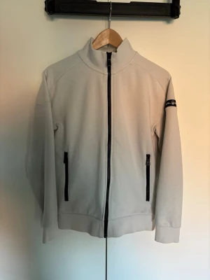 Vit ziptröja från Sail Racing - Snygg vit ziptröja från Sail Racing med hög krage och svarta detaljer. Tröjan har två dragkedjeförsedda fickor framtill och en logga på ärmen. Perfekt för en clean och sportig look. Hör av er vid frågor eller lägg ett prisförslag!