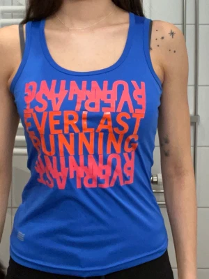 Blå linnetopp från Everlast - Snygg blå linnetopp från Everlast med stort rosa tryck framtill där det står 'EVERLAST RUNNING' i spegelvänd text. Toppen har rund halsringning och ärmlös design, perfekt för träning eller en sportig look.