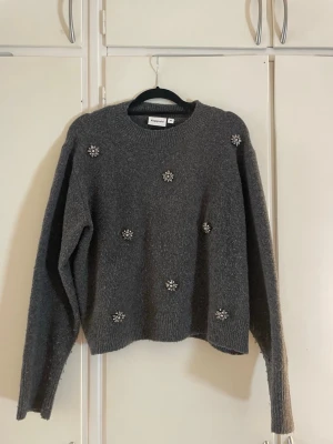 Grå stickad tröja med pärlbroderi - Mysig grå stickad tröja från Kappahl med dekorativa pärlbroderade blommor framtill. Tröjan har rund halsringning, långa ärmar och ribbade muddar vid ärmslut och nederkant. Perfekt för dig som gillar en enkel men ändå unik look.