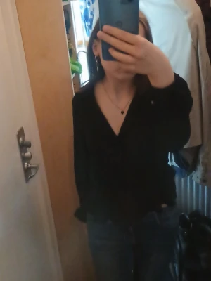 Svart blus med v-ringning - Snygg svart blus med djup v-ringning och långa ärmar. Blusen har en avslappnad passform och lätt volang nedtill, vilket ger en trendig siluett. Perfekt att matcha med jeans eller kjol för en cool look.