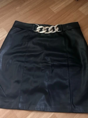 Svart kortkjol med kedjedetalj - Snygg svart kortkjol i skinnimitation med en guldfärgad kedjedetalj i midjan. Kjolen har en rak formig till rak passform 