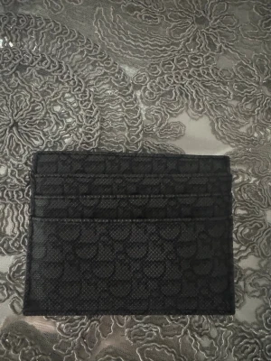 Svart kortfodral  - Snyggt svart kortfodral det klassiska mönstret. Fodralet har flera fack för kort och är gjort i tåligt konstläder. Perfekt för dig som vill ha ett stilrent och smidigt sätt att bära dina kort.