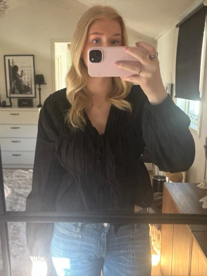 Svart blus med v-ringning och knyt - Trendig svart blus med v-ringning och knyt framtill. Blusen har långa ärmar med lätt puff och resår vid ärmslut. Från Gina