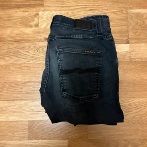 Svarta Nudie Jeans   - Snygga svarta jeans från Nudie Jeans med slim fit och klassisk femficksdesign. Jeansen har en mörk tvätt med lätt slitning på låren och bakfickor med Nudie-logga. Perfekt passform och stretchigt material för en modern look. W31 L32. Lagning i skrevet och knäna men inget man tänker på. Skriv vid frågor, pris kan diskuteras.