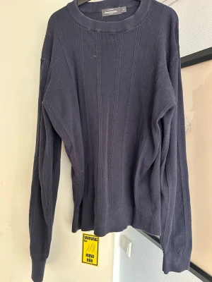 Mörkblå stickad tröja Dressmann - Mörkblå stickad tröja från Dressmann med rund halsringning och diskret ribbad struktur. Tröjan har långa ärmar och en klassisk, avslappnad passform. Perfekt för lager på lager under kyliga dagar.