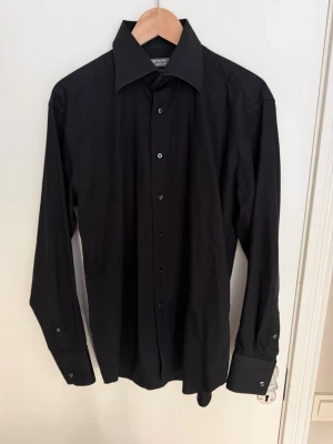 Svart skjorta med klassisk krage - Stilren svart skjorta med lång ärm, klassisk krage och knäppning framtill. Skjortan har smala manschetter med knappar och är tillverkad i ett mjukt bomullstyg som ger en clean look. Perfekt för dig som gillar minimalistisk stil.