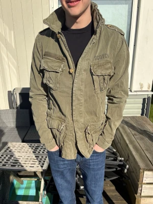 Olivgrön Scotch&soda Fieldjacka med tryck - Säljer en olivgrön Scotch&Soda Fieldjacka med hög krage och coolt tryck på ryggen. Jackan har flera fickor, axelklaffar och knappar framtill. Perfekt för dig som gillar streetwear och vill sticka ut lite extra.