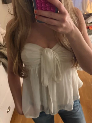 Vit bandeau topp med stor rosett - Supersöt vit bandeau topp med stor rosett framtill. Toppen är axelbandslös och har ett luftigt, lätt material som ger en romantisk känsla. Perfekt att styla med jeans eller kjol för en trendig look.