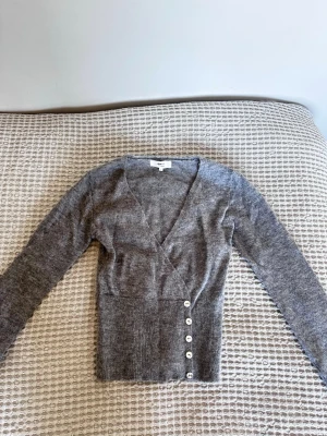 Trendig Gina tröja - Mysig grå stickad tröja från Gina Tricot i alpackablandning. Tröjan har djup v-ringning, långa ärmar och ribbad midja med dekorativa vita knappar på sidan. Perfekt för lager på lager och en chill look. Använd 1 gång❣️