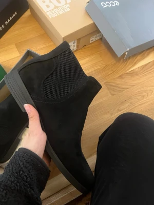 Svarta Chelsea boots från din sko  - Säljer ett par stilrena svarta Chelsea boots från din sko  i mocka med elastiska paneler på sidorna och låg klack. Skorna har en klassisk siluett och är perfekta för dig som gillar enkel och tidlös design. Sula i gummi för bra grepp. Helt nya och oanvänd. 