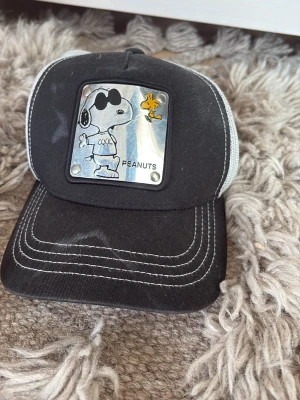 Svart Peanuts keps från Goorin Bros med Snoopy - Säljer en svart keps med vit mesh på sidorna och en metallbricka framtill med Snoopy och Woodstock från Peanuts. Kepsen har vita kontrastsömmar och justerbar baksida. Perfekt för dig som gillar coola och lekfulla accessoarer.